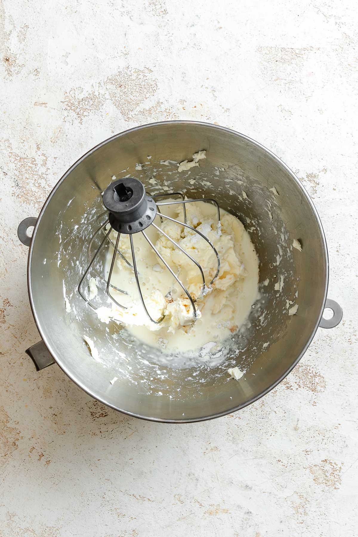 Mascarpone, cream, vanilla, and powdered sugar partially mixed together in a stand mixer bowl with the whisk attachment.