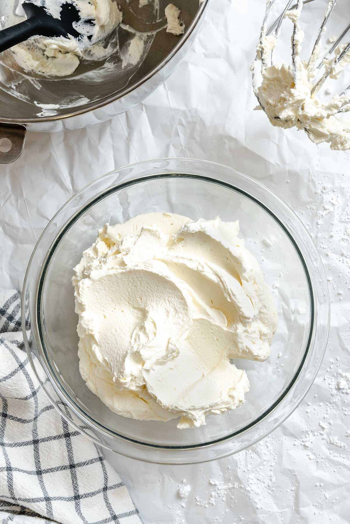 Finished mascarpone whipped cream transferred to a clear glass bowl, with spatula and stand mixer bowl in the background.