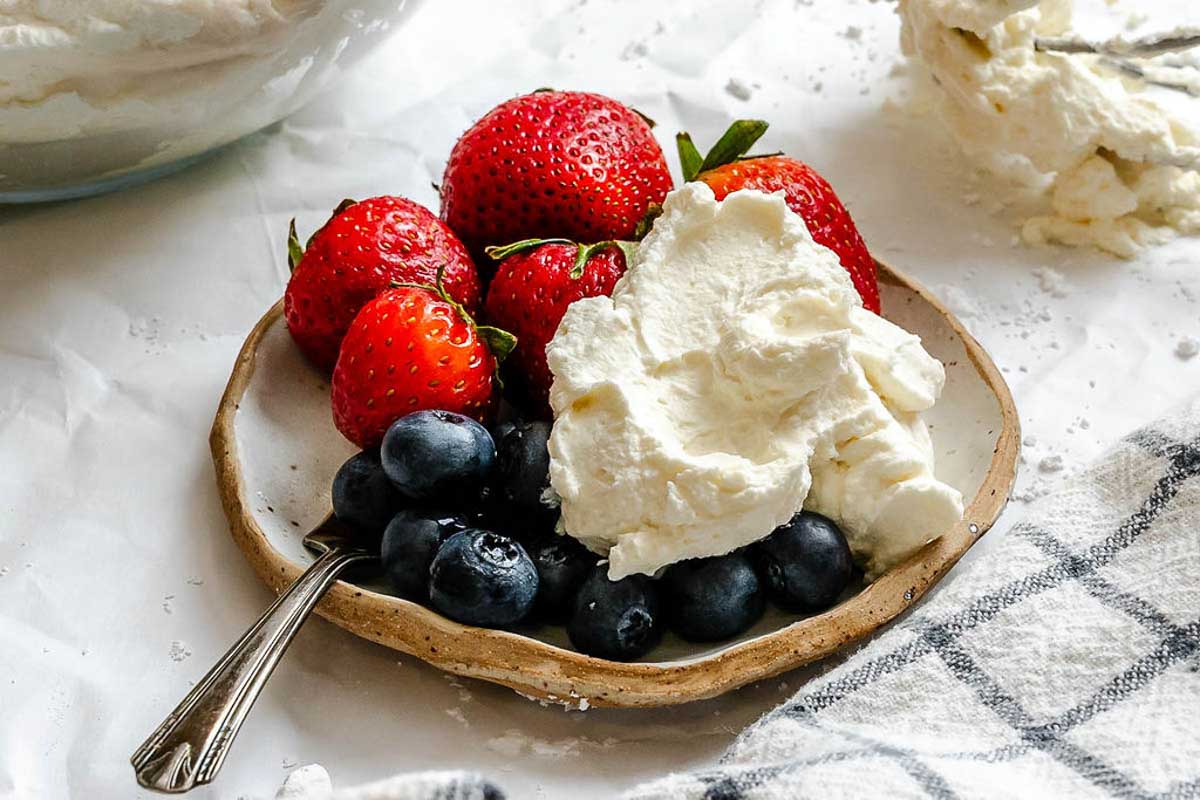 Small plate of fresh strawberries and blueberries topped with a generous scoop of mascarpone whipped cream, with whisk attachment and mixing bowl in the background.