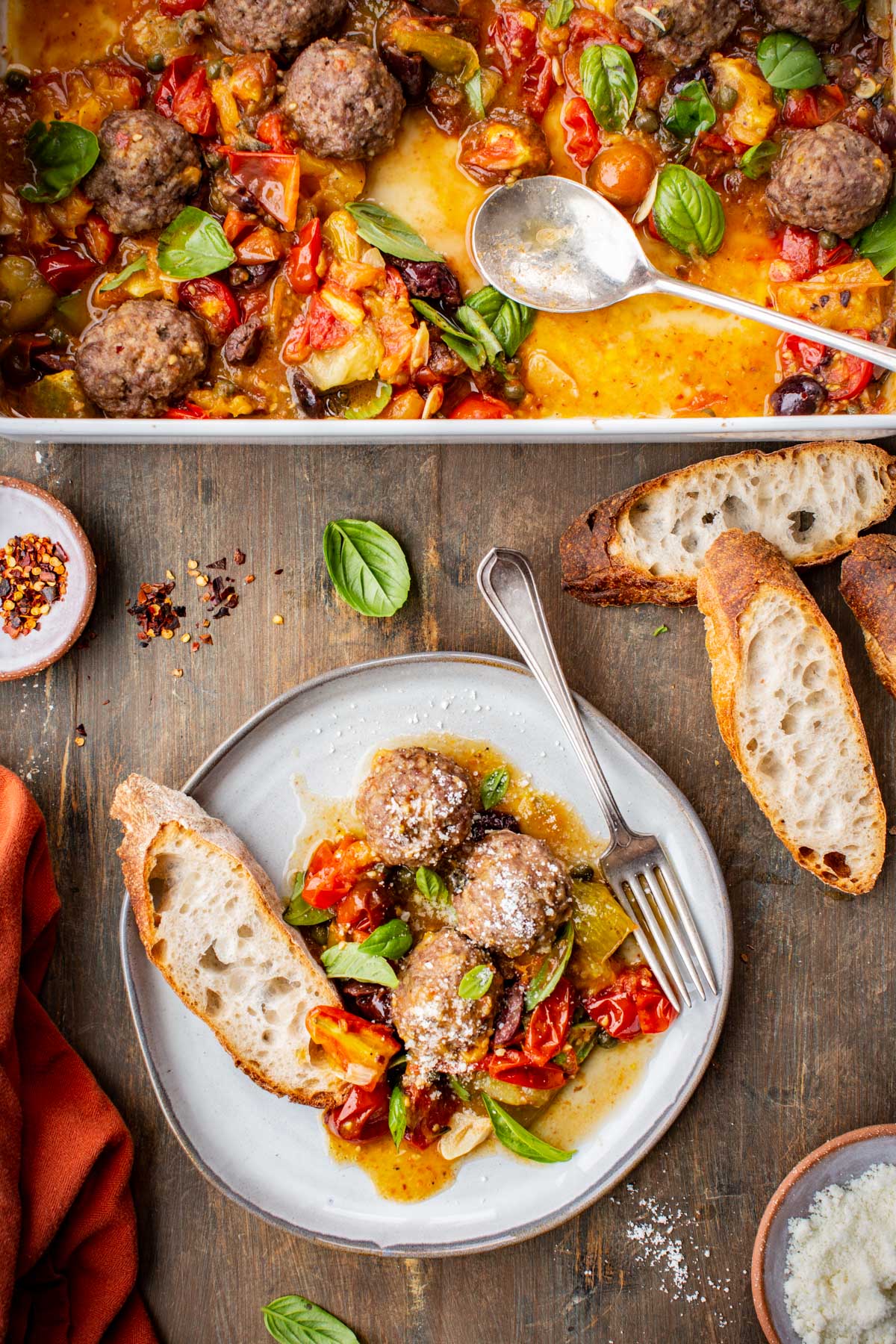 Plate of meatballs with puttanesca sauce, sprinkled with grated cheese and served with crusty bread, next to the casserole dish.