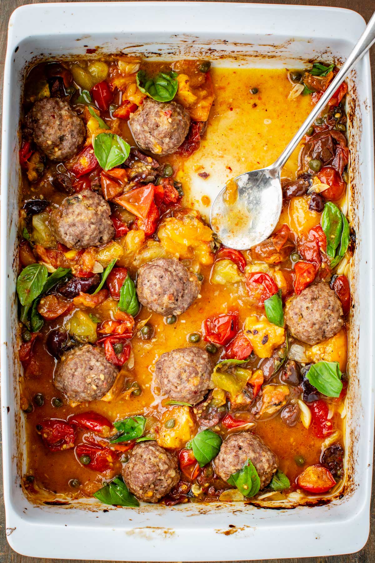Partially eaten casserole dish of roasted meatballs with tomato sauce, olives, and basil, with a spoon resting inside.