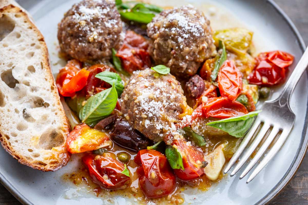 Close-up of three meatballs with roasted tomatoes, olives, and basil, sprinkled with cheese, served with bread on the side.