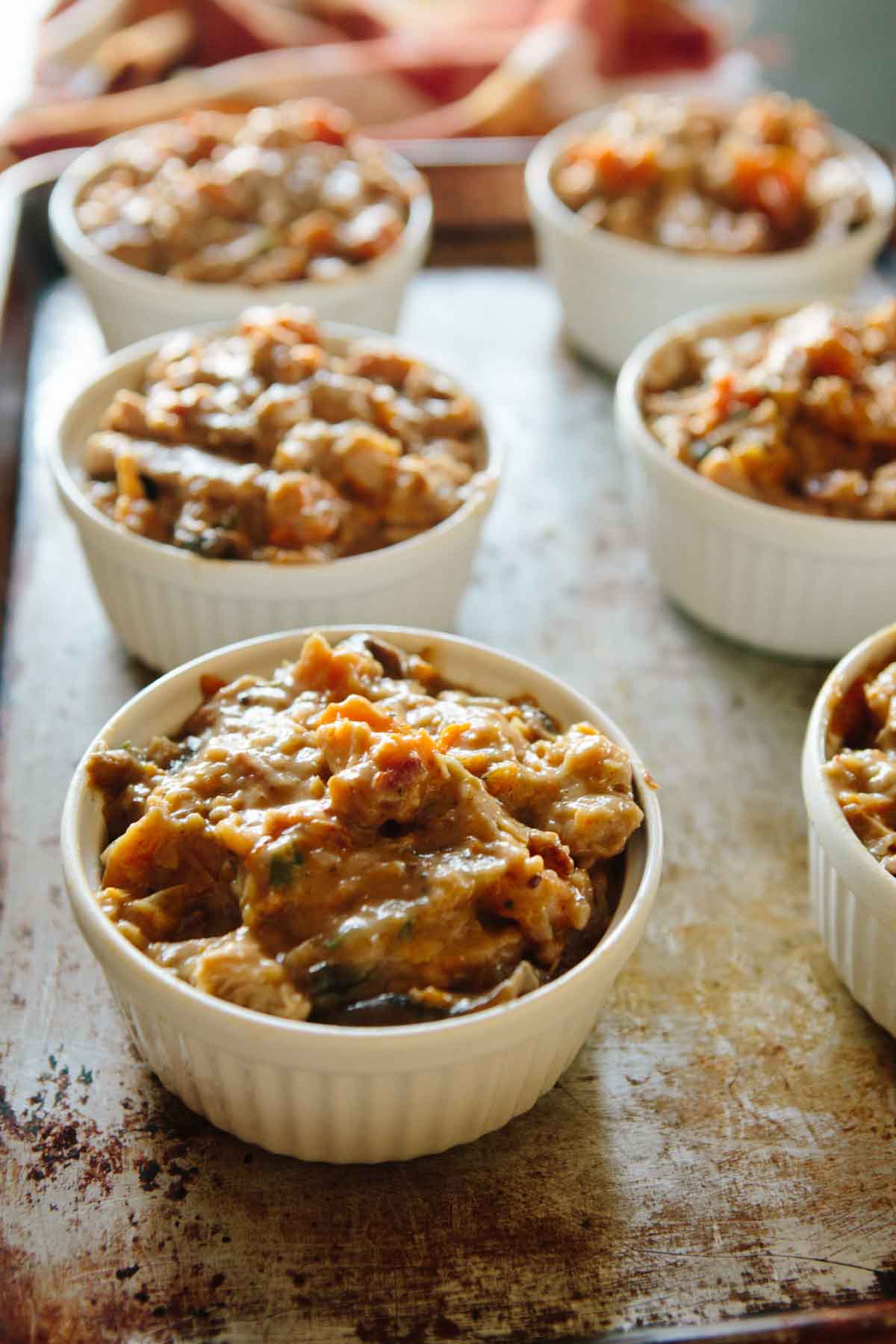 White ramekins filled with hearty turkey pot pie filling made with butternut squash, mushrooms, onions, and bacon, set on a baking sheet before being topped with crust.