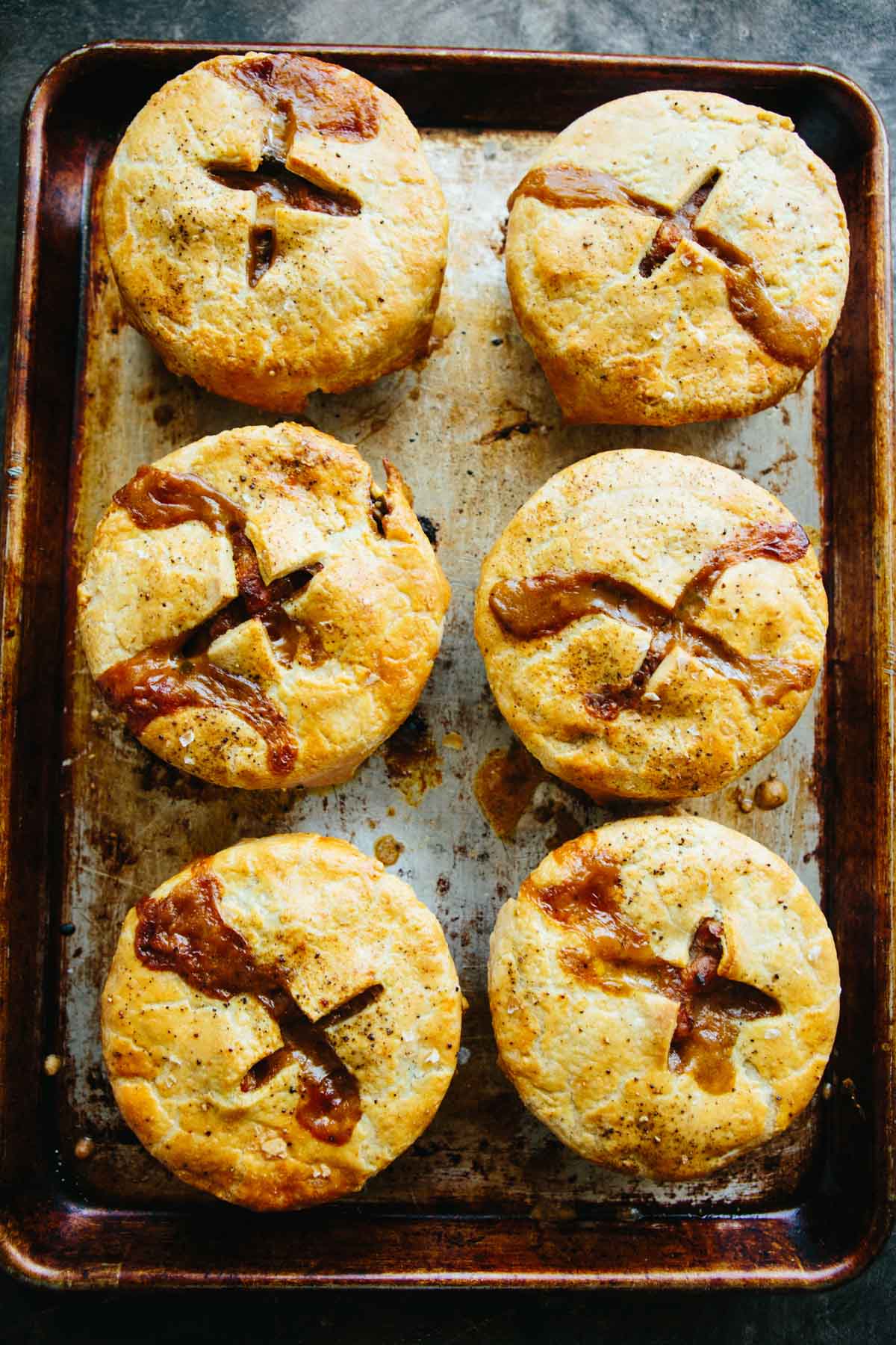 Overhead of six mini turkey pot pies on a baking sheet, crusts deeply golden with rustic cracks showing filling beneath.