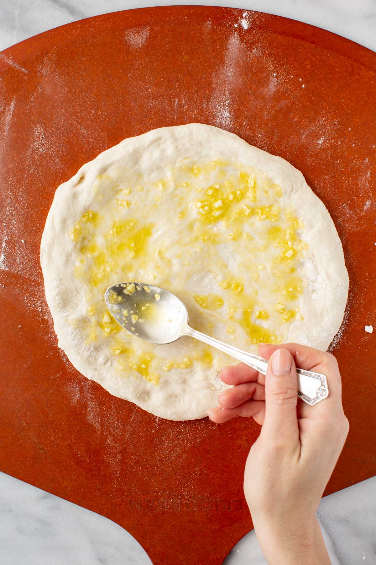 Brushing garlic oil - A spoon spreading garlic oil over stretched pizza dough on a floured pizza peel before adding cheese.