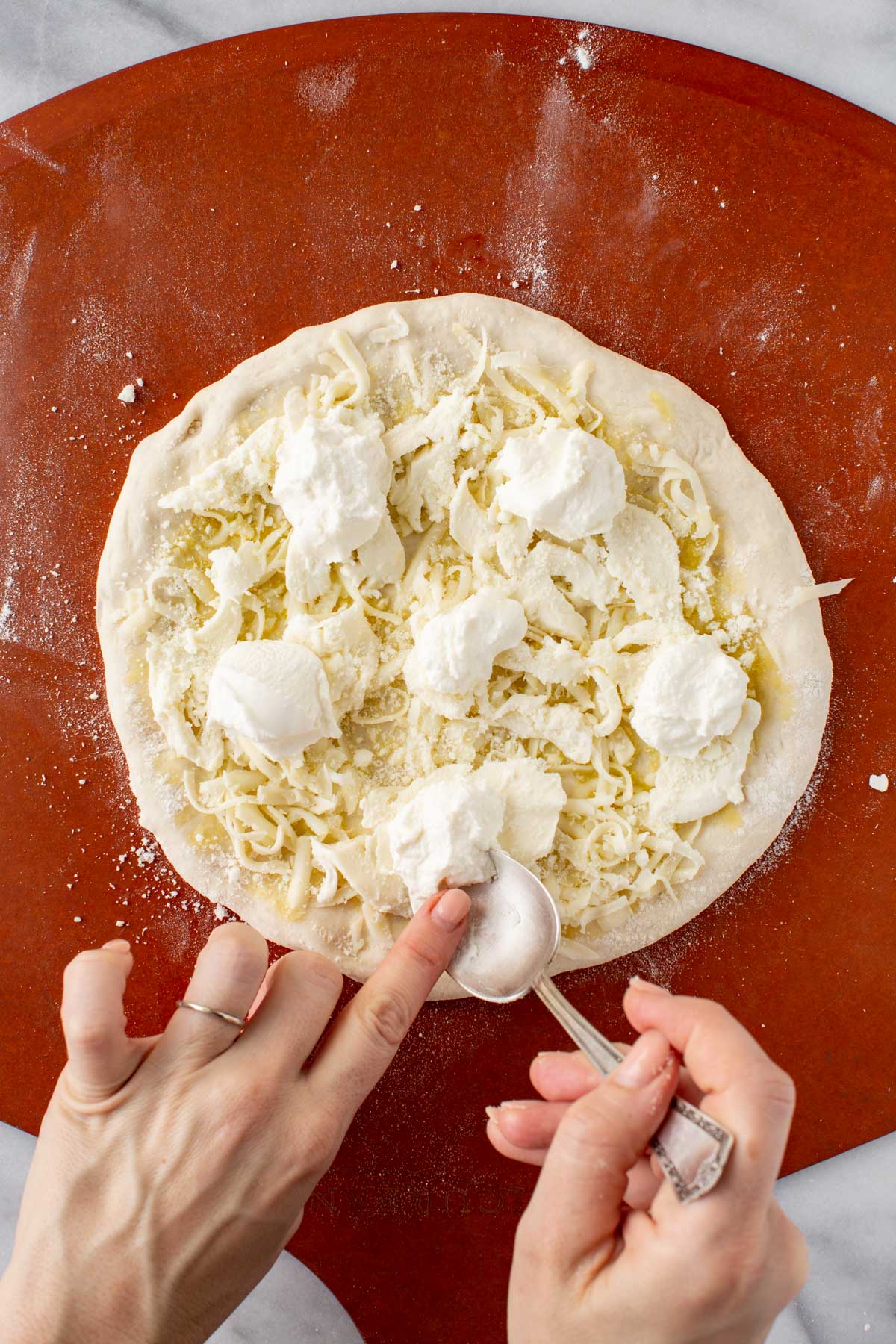 Adding ricotta - Hands using a spoon to place dollops of ricotta cheese over a pizza already topped with mozzarella.