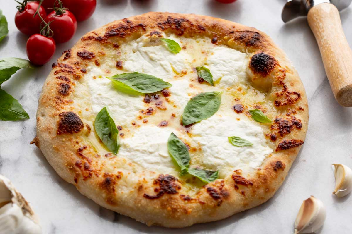 Styled whole pizza – Pizza bianca garnished with basil, photographed with fresh tomatoes, garlic, and a pizza cutter for serving.