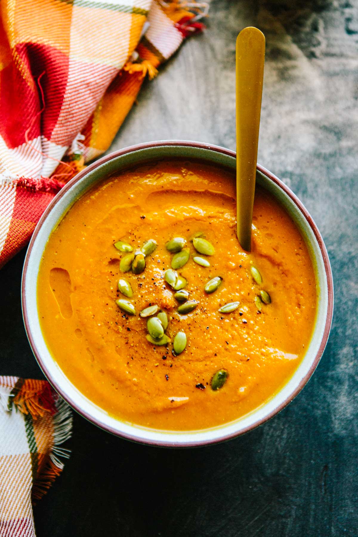 Overhead view of a bowl filled with smooth pumpkin soup, garnished with pumpkin seeds and black pepper, set beside a colorful plaid napkin.