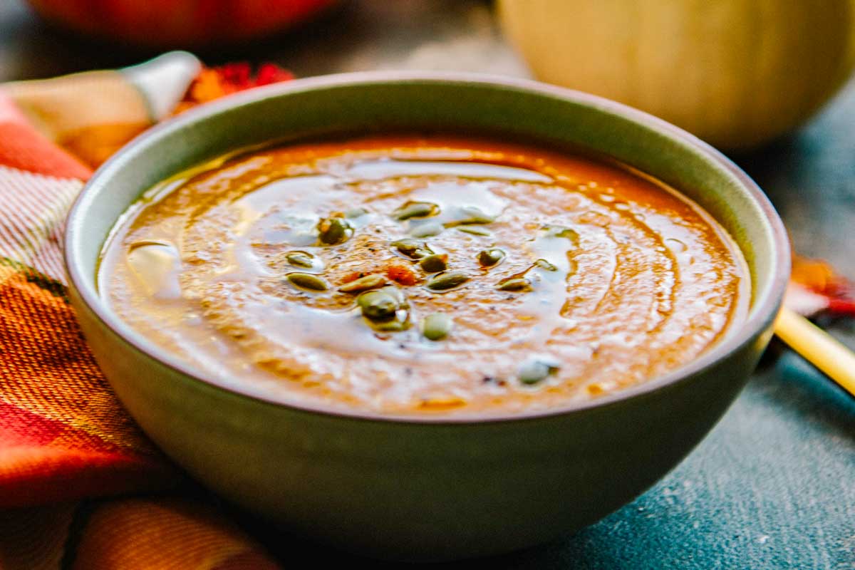 Side view of a rustic bowl of pumpkin soup, swirled with oil and topped with seeds, photographed with warm autumn tones and fabric nearby.