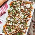 Overhead view of a large rectangular serving platter filled with roasted butternut squash, goat cheese, hazelnuts, and herbs, with whole butternut squash and a red-striped cloth in the background.