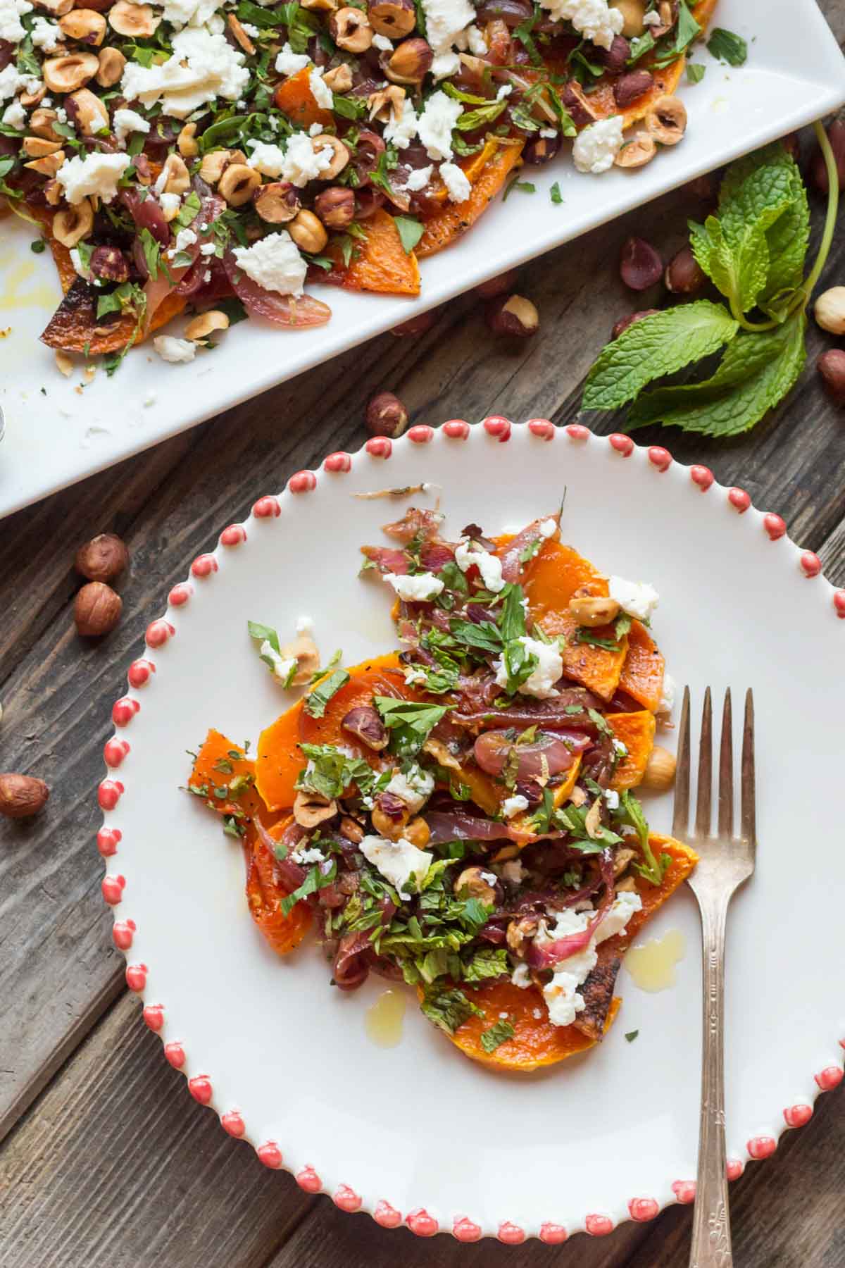 Overhead shot of a white plate with scalloped red trim filled with roasted butternut squash topped with caramelized red onions, crumbled goat cheese, toasted hazelnuts, and fresh herbs, with a fork on the side.