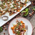 Overhead view of a large rectangular white platter heaped with roasted butternut squash, onions, goat cheese, hazelnuts, and herbs, shown next to a smaller plated serving and scattered hazelnuts and mint leaves.