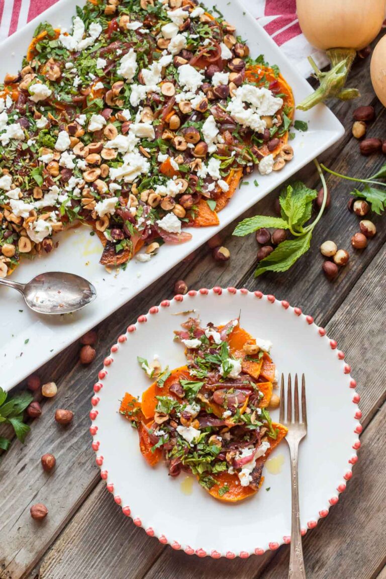 Overhead view of a large rectangular white platter heaped with roasted butternut squash, onions, goat cheese, hazelnuts, and herbs, shown next to a smaller plated serving and scattered hazelnuts and mint leaves.