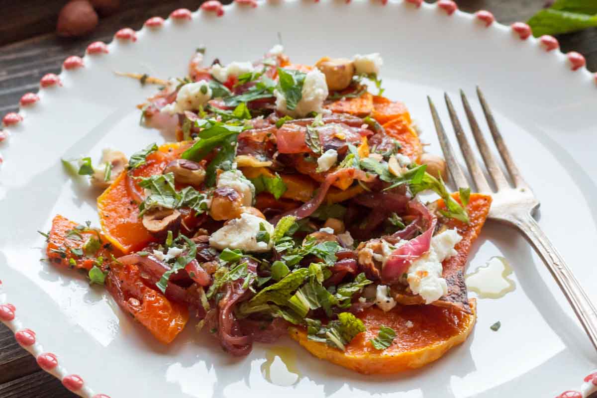 Close-up of a plated serving of roasted butternut squash with onions, goat cheese, hazelnuts, and fresh herbs, highlighting the textures and drizzled olive oil.