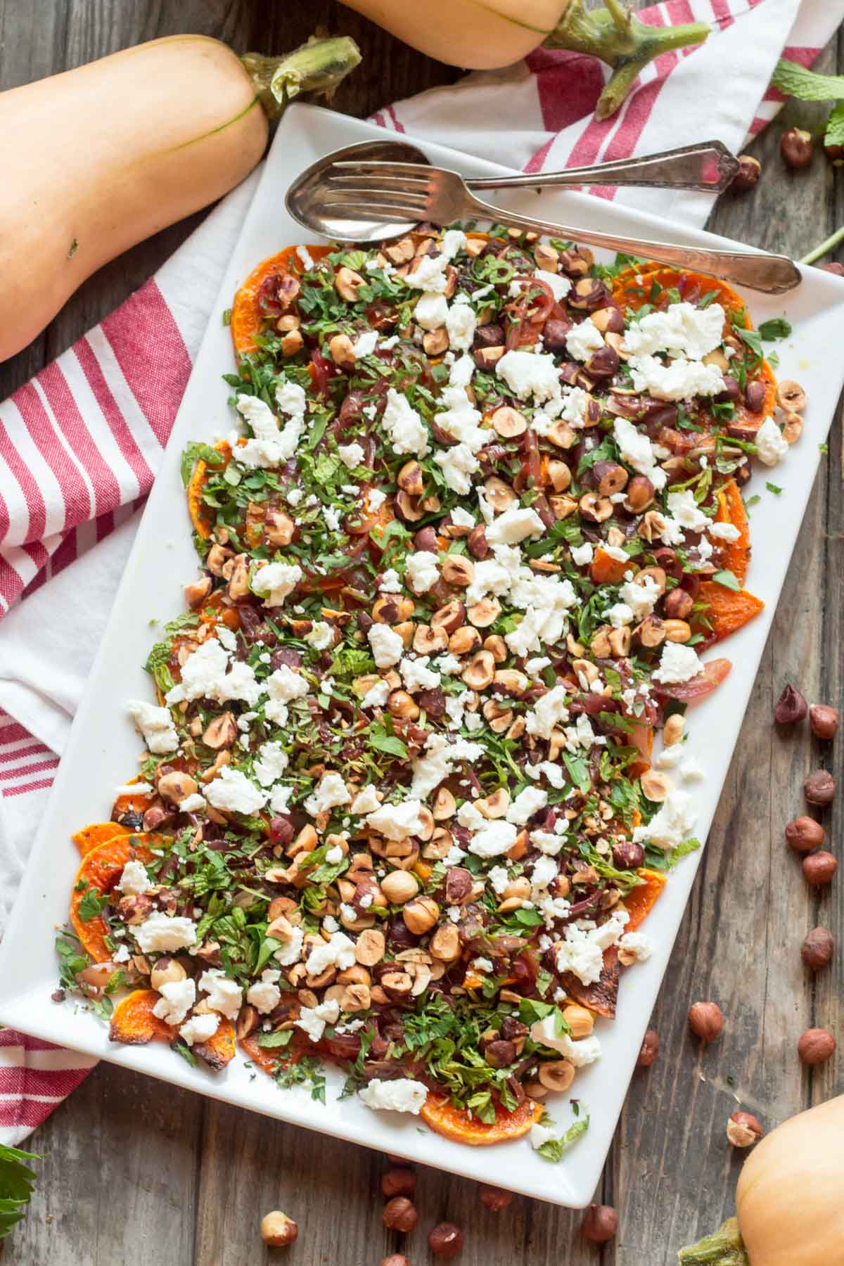 Overhead view of a large rectangular serving platter filled with roasted butternut squash, goat cheese, hazelnuts, and herbs, with whole butternut squash and a red-striped cloth in the background.