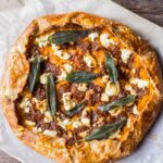 Whole pumpkin and goat cheese galette on parchment paper with deeply golden crust, browned onion filling, scattered sage leaves, and crumbles of goat cheese.