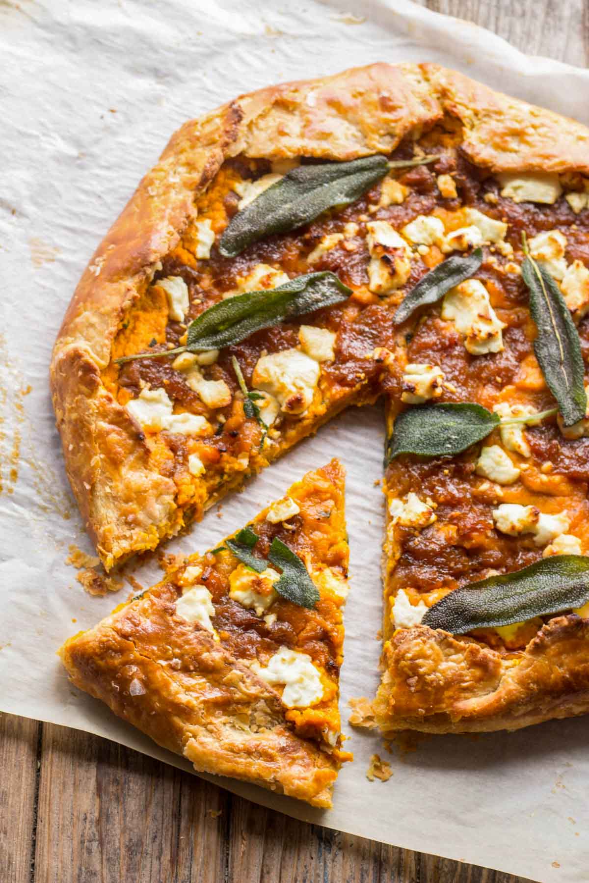 A rustic pumpkin and goat cheese galette on parchment paper with golden crust, caramelized onions, goat cheese crumbles, and crispy sage leaves, with one slice cut out.