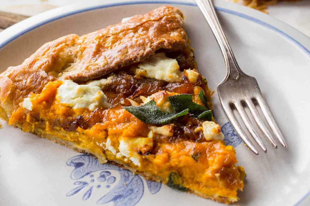 A plated slice of pumpkin and goat cheese galette on a white dish with a fork, highlighting the flaky crust, bright orange pumpkin filling, caramelized onions, and goat cheese.