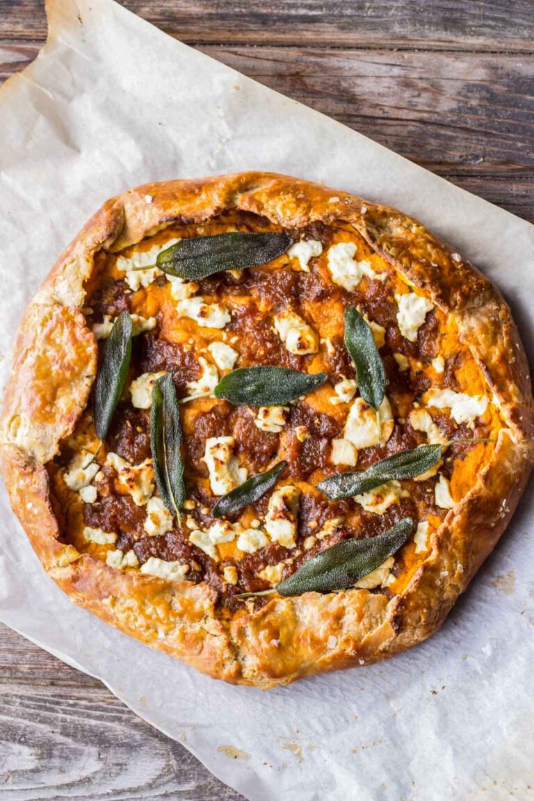 Whole pumpkin and goat cheese galette on parchment paper with deeply golden crust, browned onion filling, scattered sage leaves, and crumbles of goat cheese.