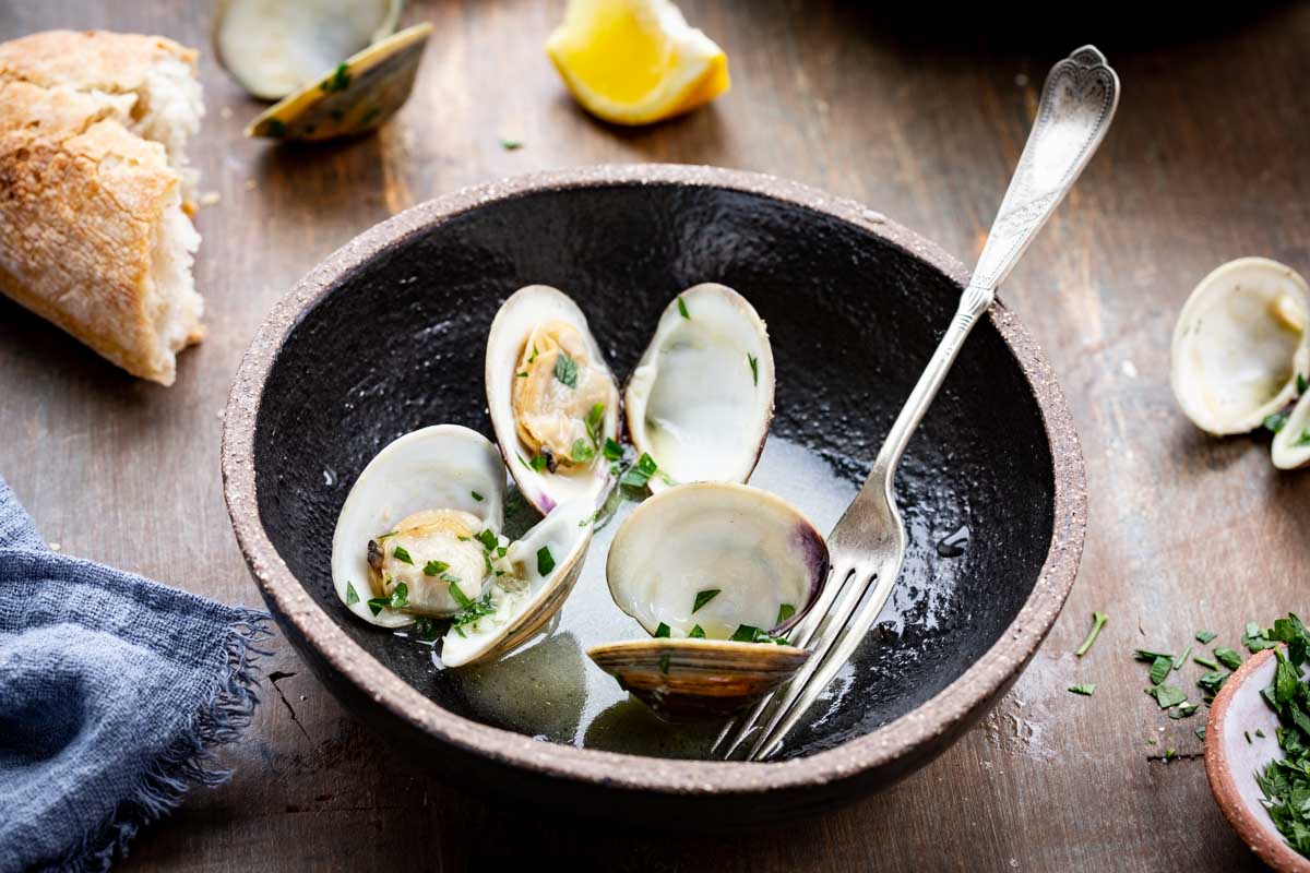 A close-up of a small black bowl holding several opened clams in broth, garnished with parsley, with a fork resting inside and crusty bread in the background.