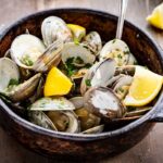 Steamed clams in their shells served in a dark ceramic bowl, topped with parsley and lemon wedges, with clam shells and fresh herbs scattered on the wooden surface around it.
