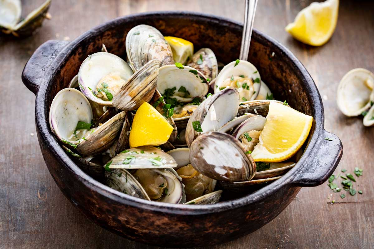 Steamed clams in their shells served in a dark ceramic bowl, topped with parsley and lemon wedges, with clam shells and fresh herbs scattered on the wooden surface around it.
