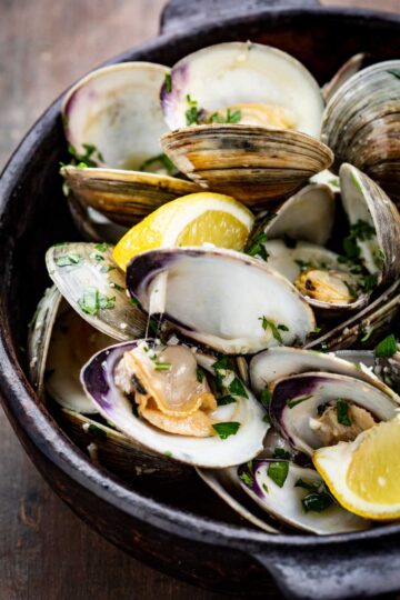 A detailed view of opened clams in their shells, sprinkled with parsley and served with lemon wedges in a rustic black bowl.