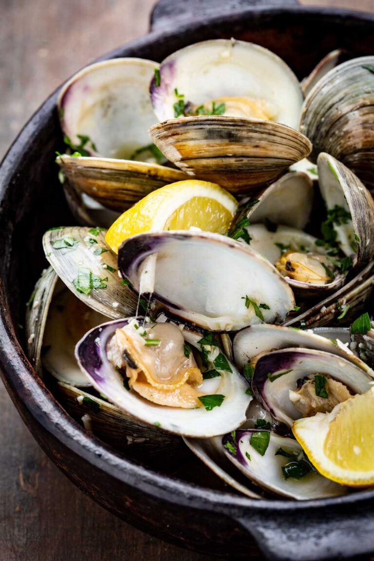 A detailed view of opened clams in their shells, sprinkled with parsley and served with lemon wedges in a rustic black bowl.