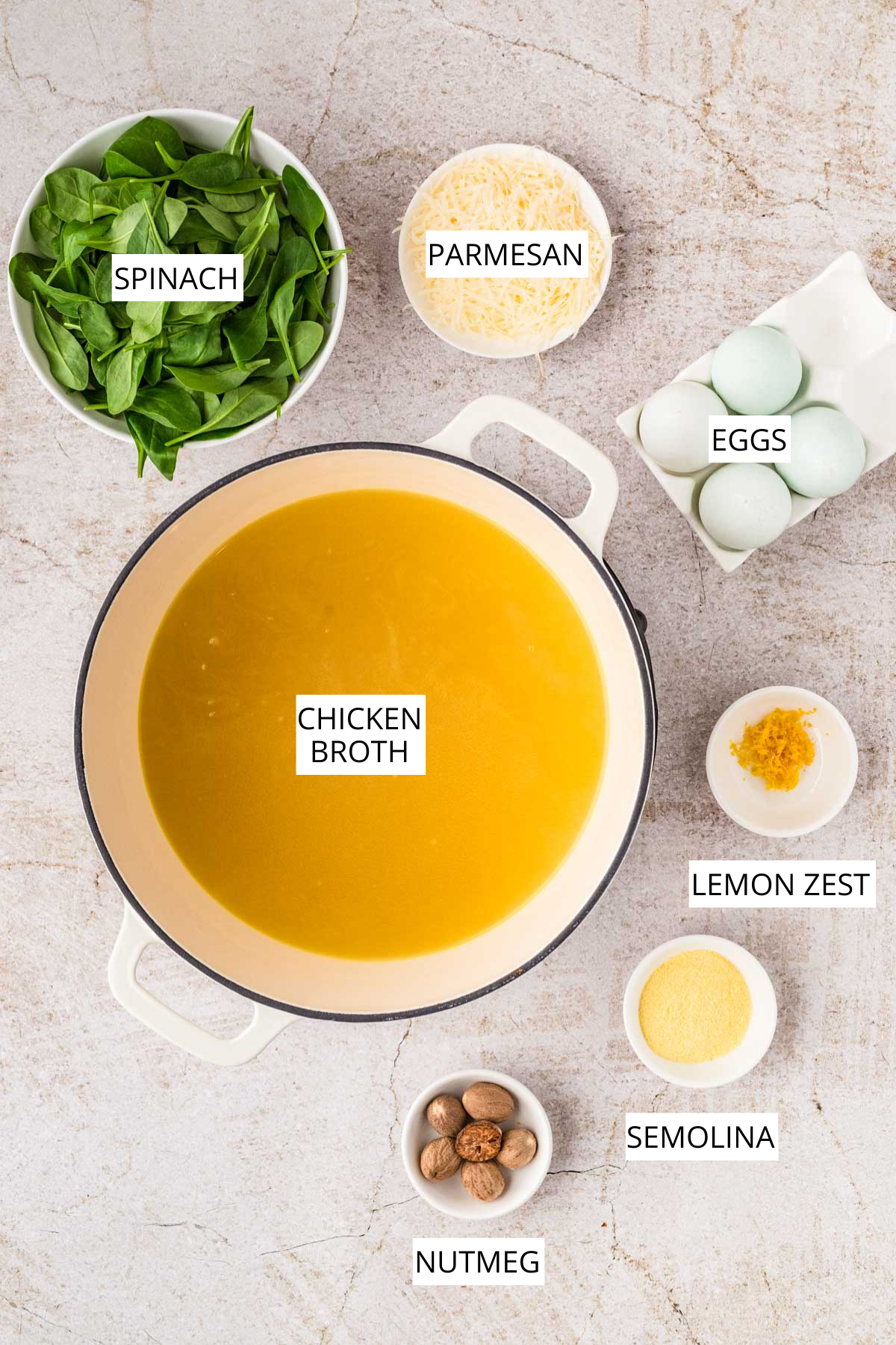 Overhead shot of the main ingredients for stracciatella soup laid out: a pot of chicken broth, fresh spinach, Parmesan, eggs, lemon zest, semolina, and nutmeg.