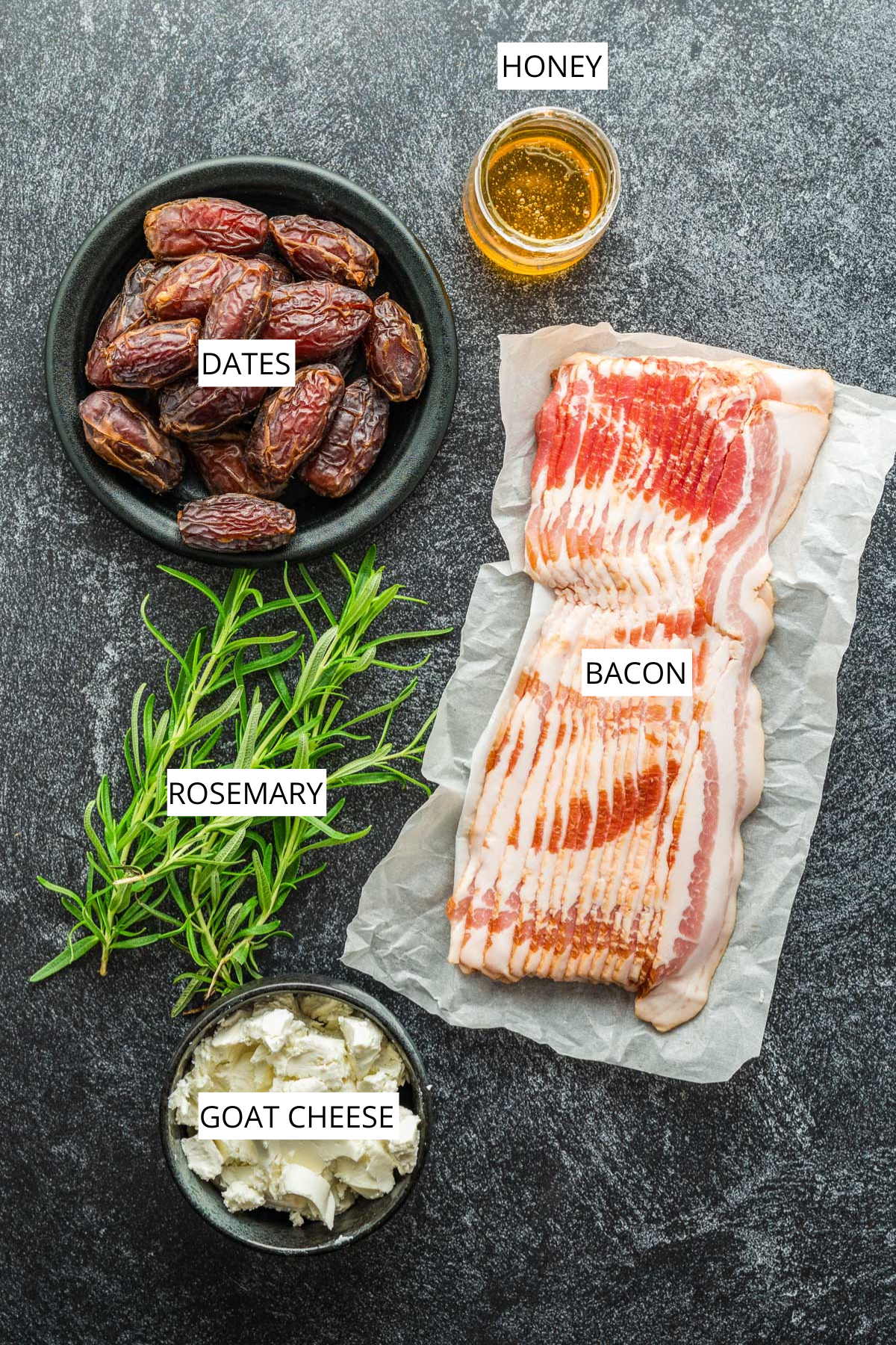 Overhead image of ingredients for bacon-wrapped stuffed dates showing dates, bacon, goat cheese, honey, and rosemary labeled on a dark surface.