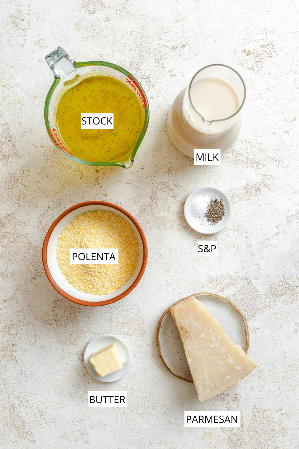 Overhead shot of measured ingredients for creamy parmesan polenta, including stock, milk, polenta cornmeal, butter, parmesan cheese, salt, and pepper arranged on a light surface.