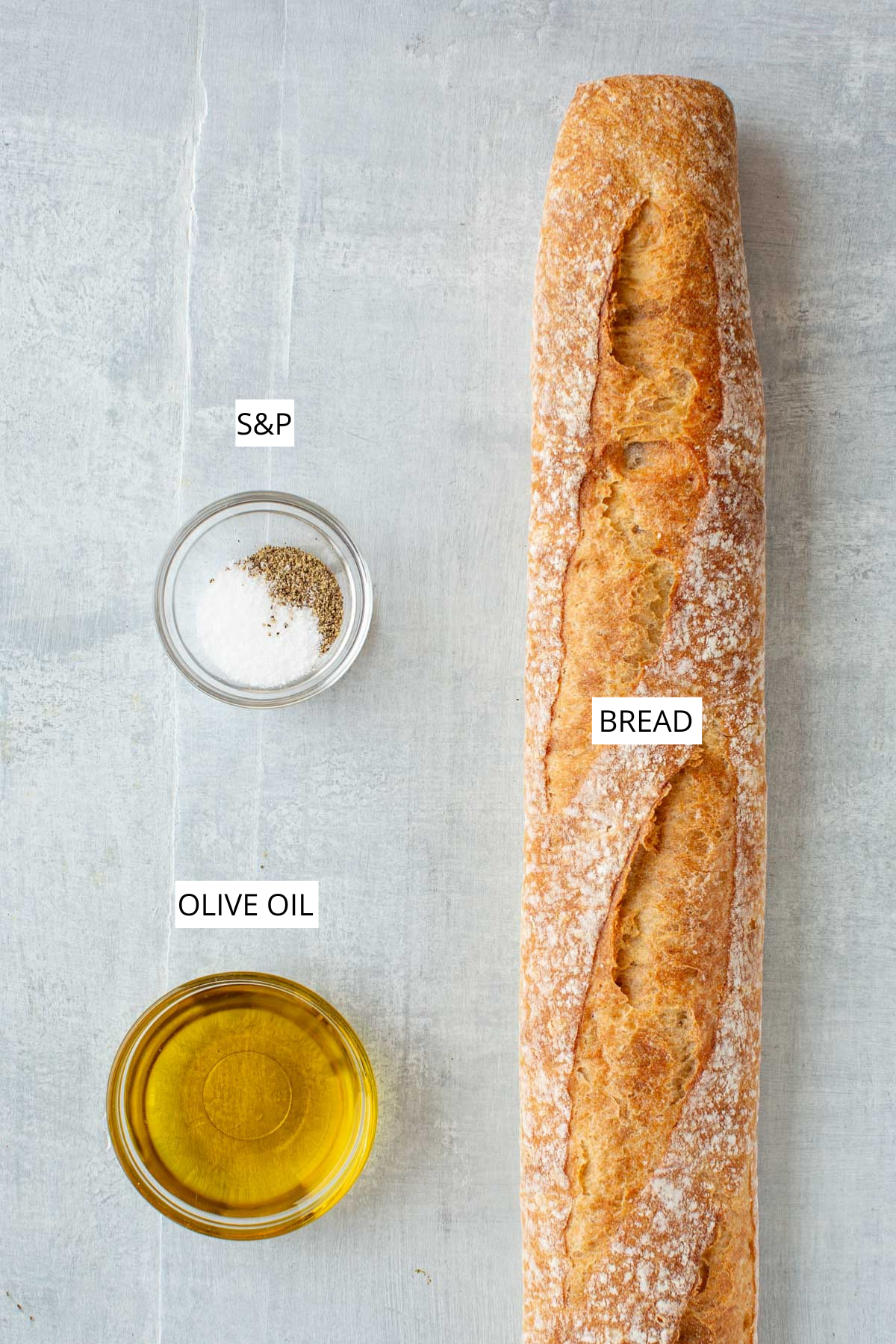 Overhead image of the three ingredients needed for homemade crostini: a crusty baguette, olive oil, and a small bowl of salt and pepper on a light gray surface.