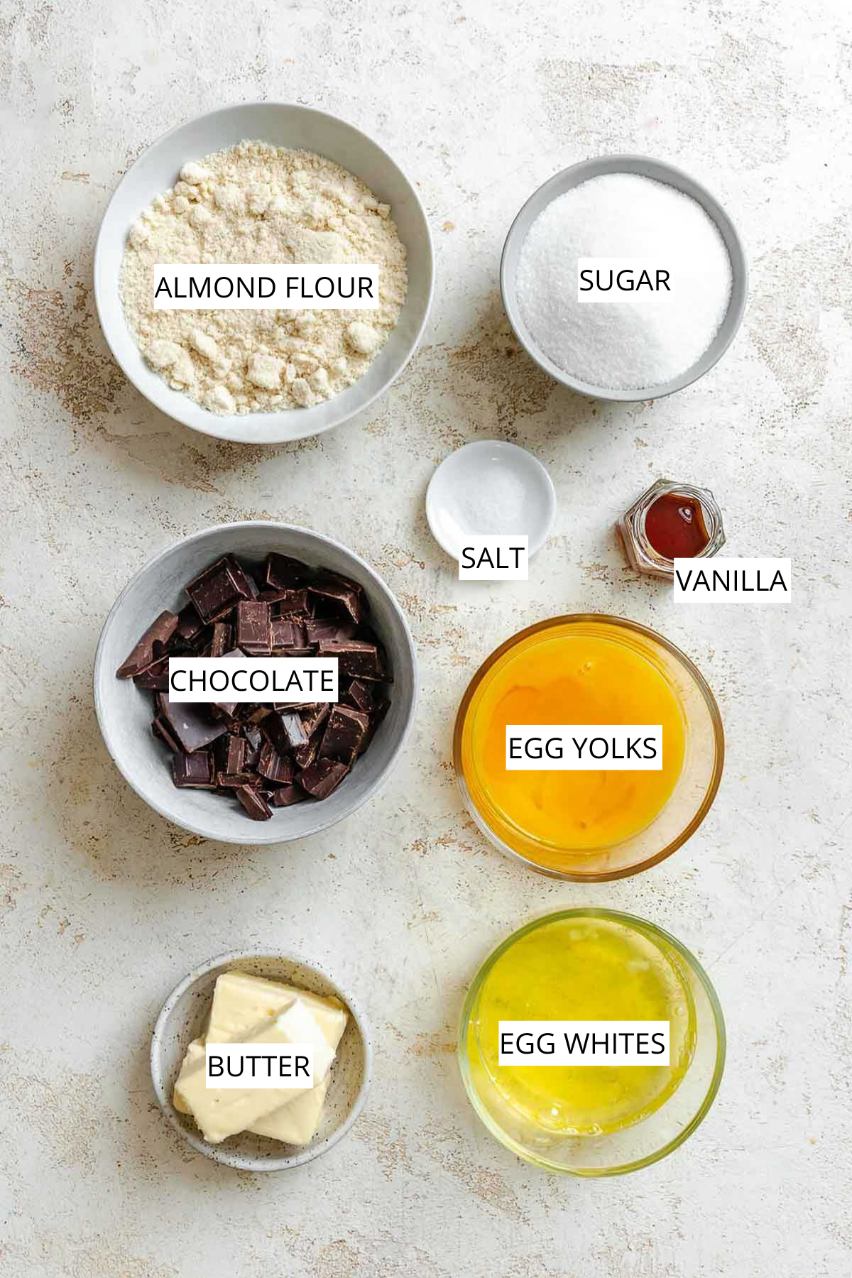 Overhead view of measured ingredients for Torta Caprese, including bowls of almond flour, sugar, chopped chocolate, butter, egg yolks, egg whites, vanilla, and salt arranged neatly on a light surface.
