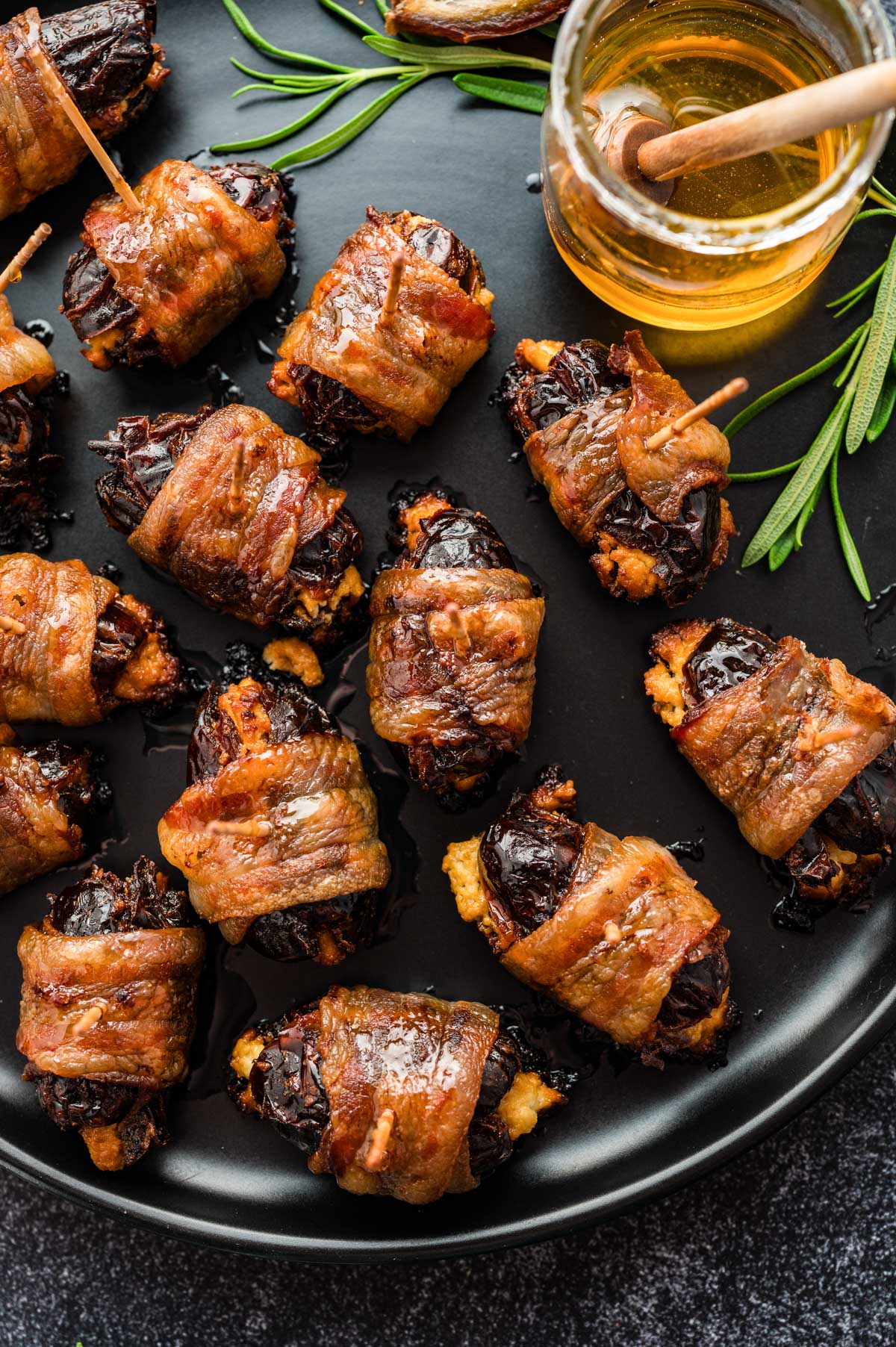 Plated bacon-wrapped dates with rosemary sprigs and a jar of honey for serving.