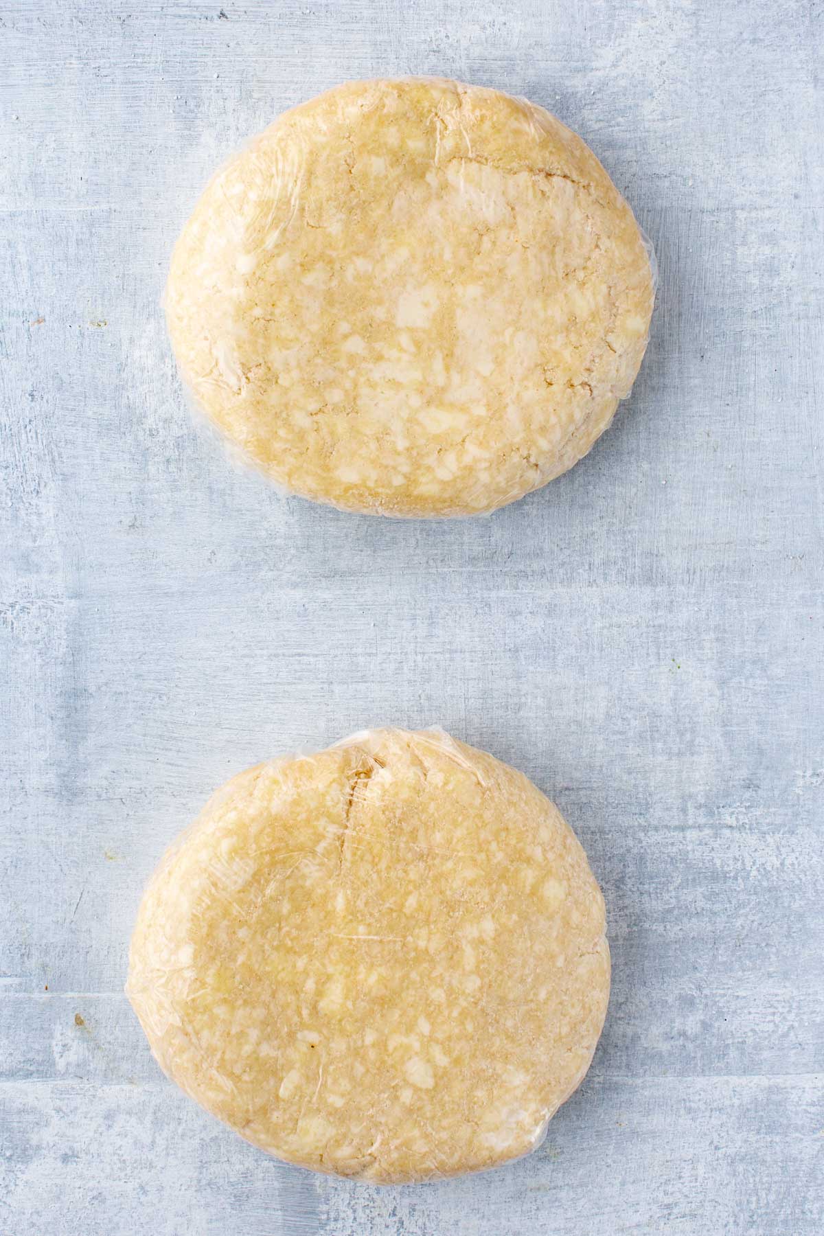Two discs of buttermilk pie dough wrapped in plastic wrap on a light blue surface, ready to chill.