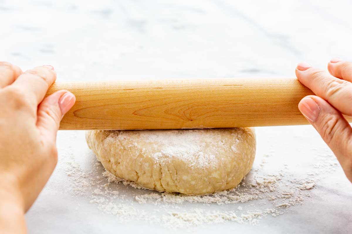Hands rolling out pie dough with a wooden rolling pin on a lightly floured surface.