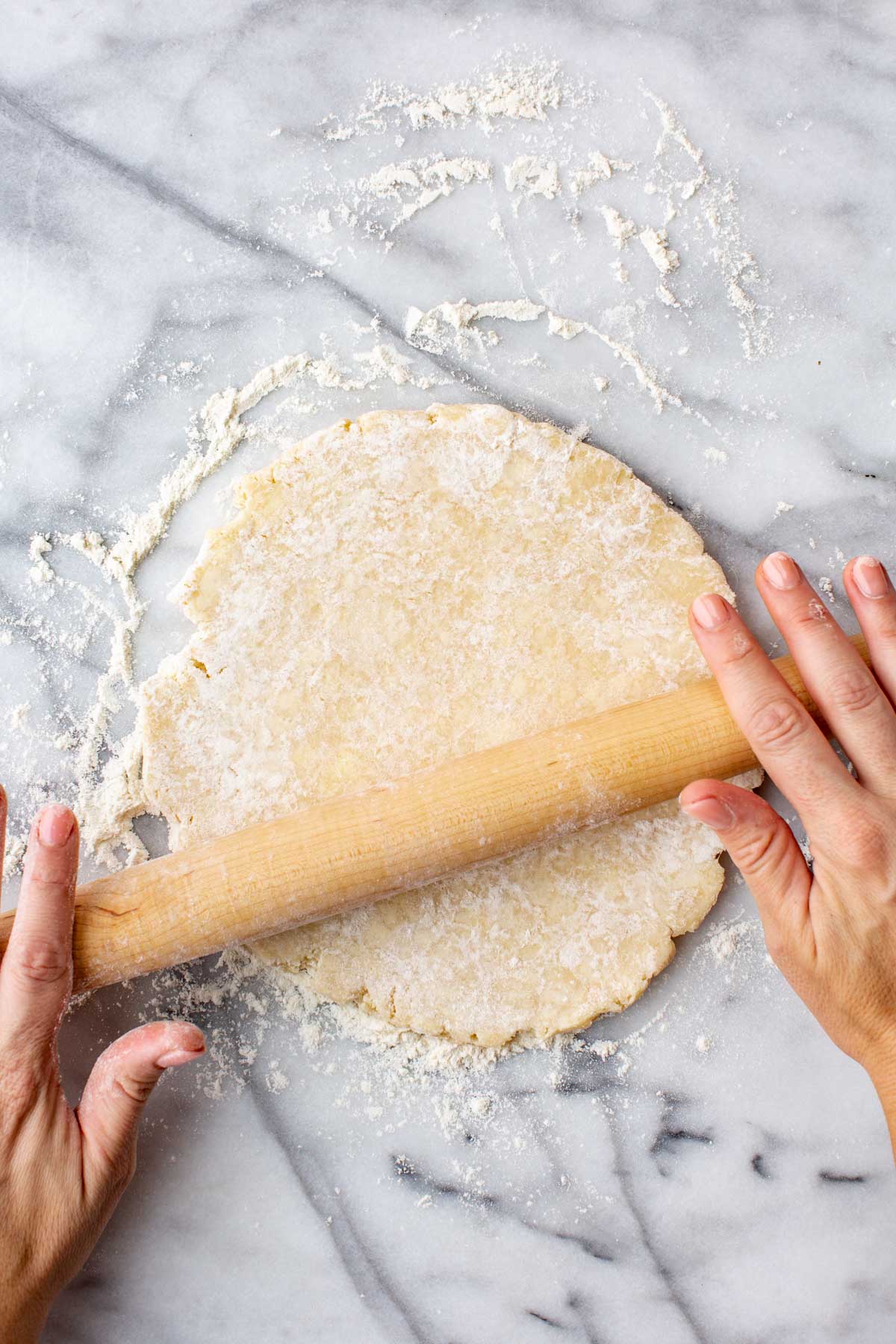 Overhead view of pie dough being rolled out evenly on a marble surface dusted with flour.