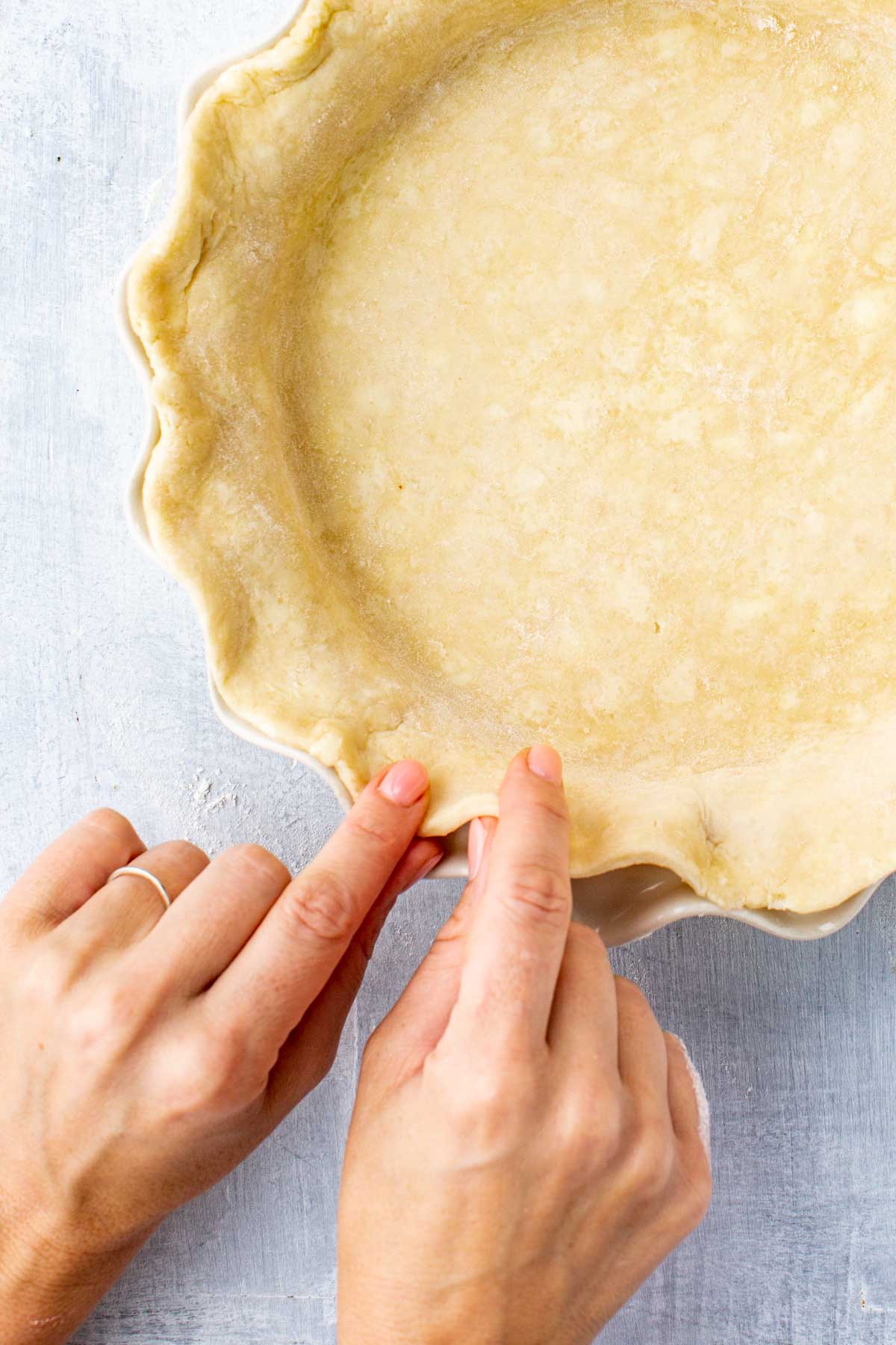 Hands crimping the edge of an unbaked pie crust in a ceramic pie dish.
