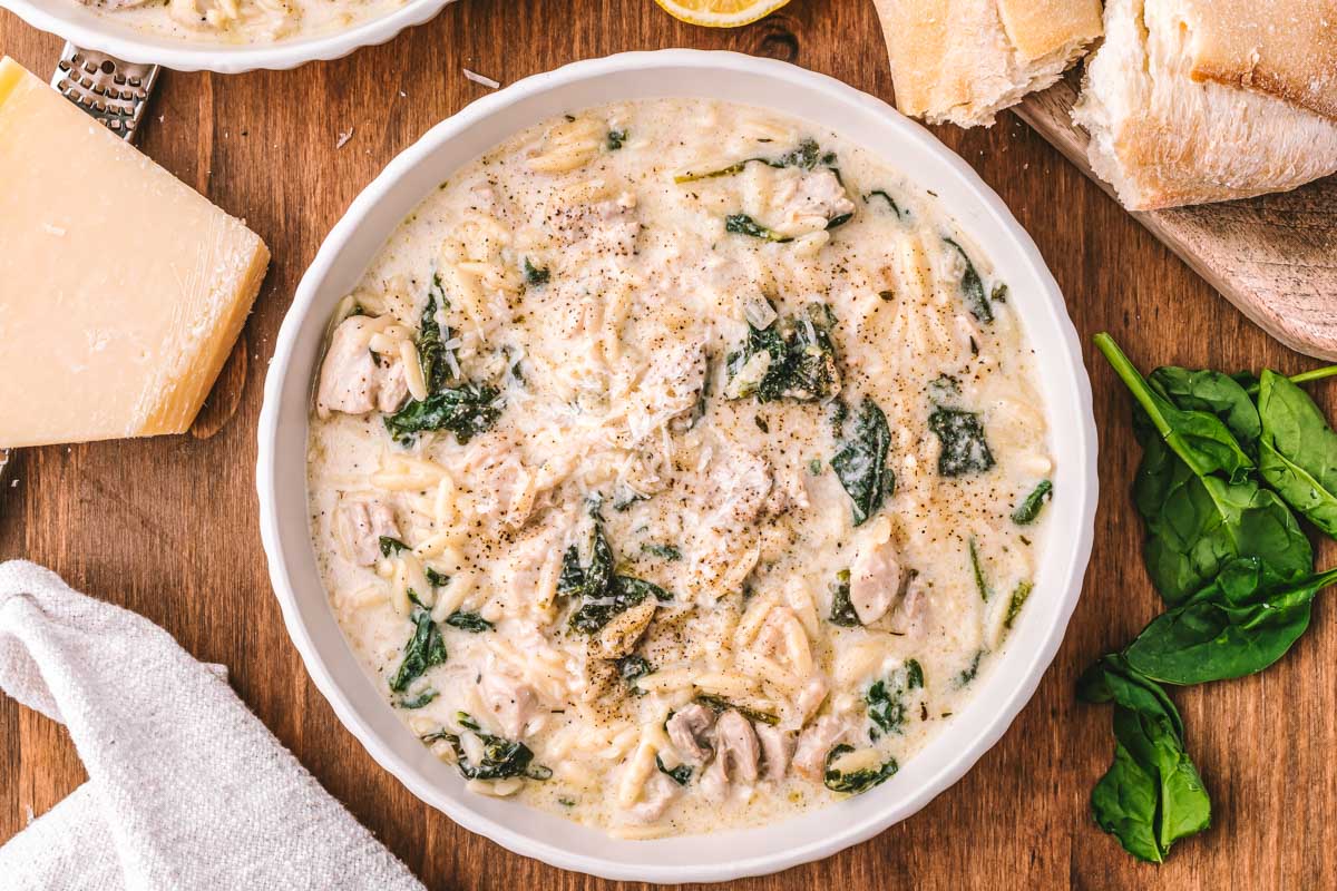 Chicken Florentine soup served in a white bowl alongside bread, lemon wedges, Parmesan cheese, and fresh spinach leaves.