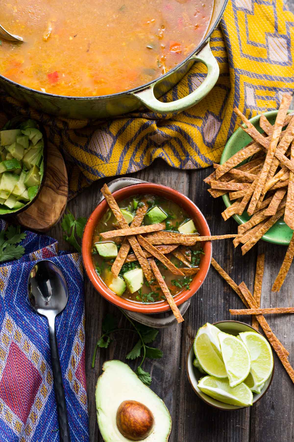 A rustic bowl of chicken tortilla soup topped with crispy tortilla strips and chunks of avocado, surrounded by lime wedges, extra strips, and a Dutch oven full of soup.
