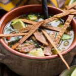 Close-up of chicken tortilla soup in a clay bowl, garnished with golden tortilla strips, avocado cubes, and fresh cilantro in a flavorful broth.