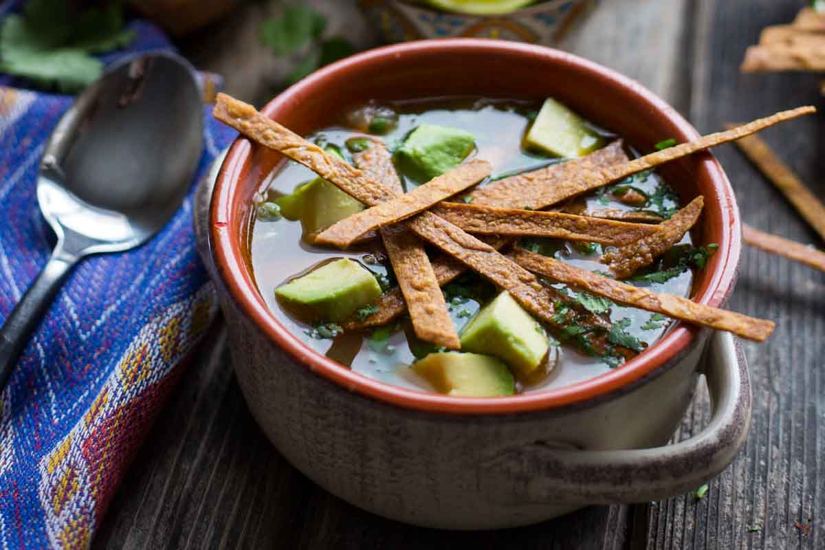 A warm ceramic bowl of chicken tortilla soup topped with avocado chunks and crunchy tortilla strips, set next to a spoon on a colorful woven napkin.