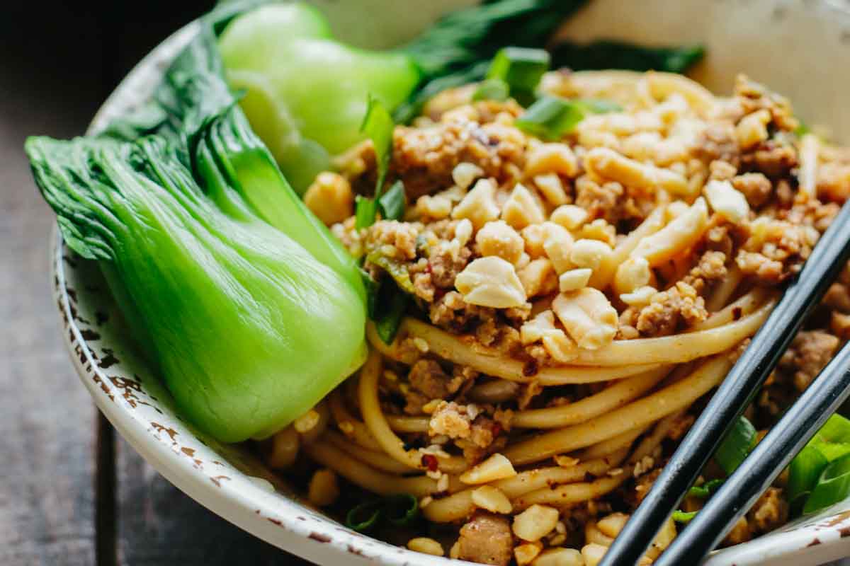 Close-up of Dan Dan noodles in a ceramic bowl topped with ground pork, crushed peanuts, scallions, and bok choy.