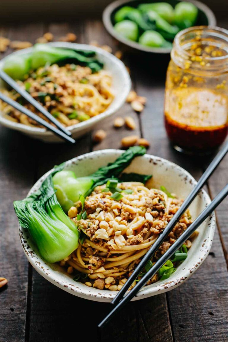 Two bowls of Dan Dan noodles with chili oil jar and bok choy on a rustic wooden table.