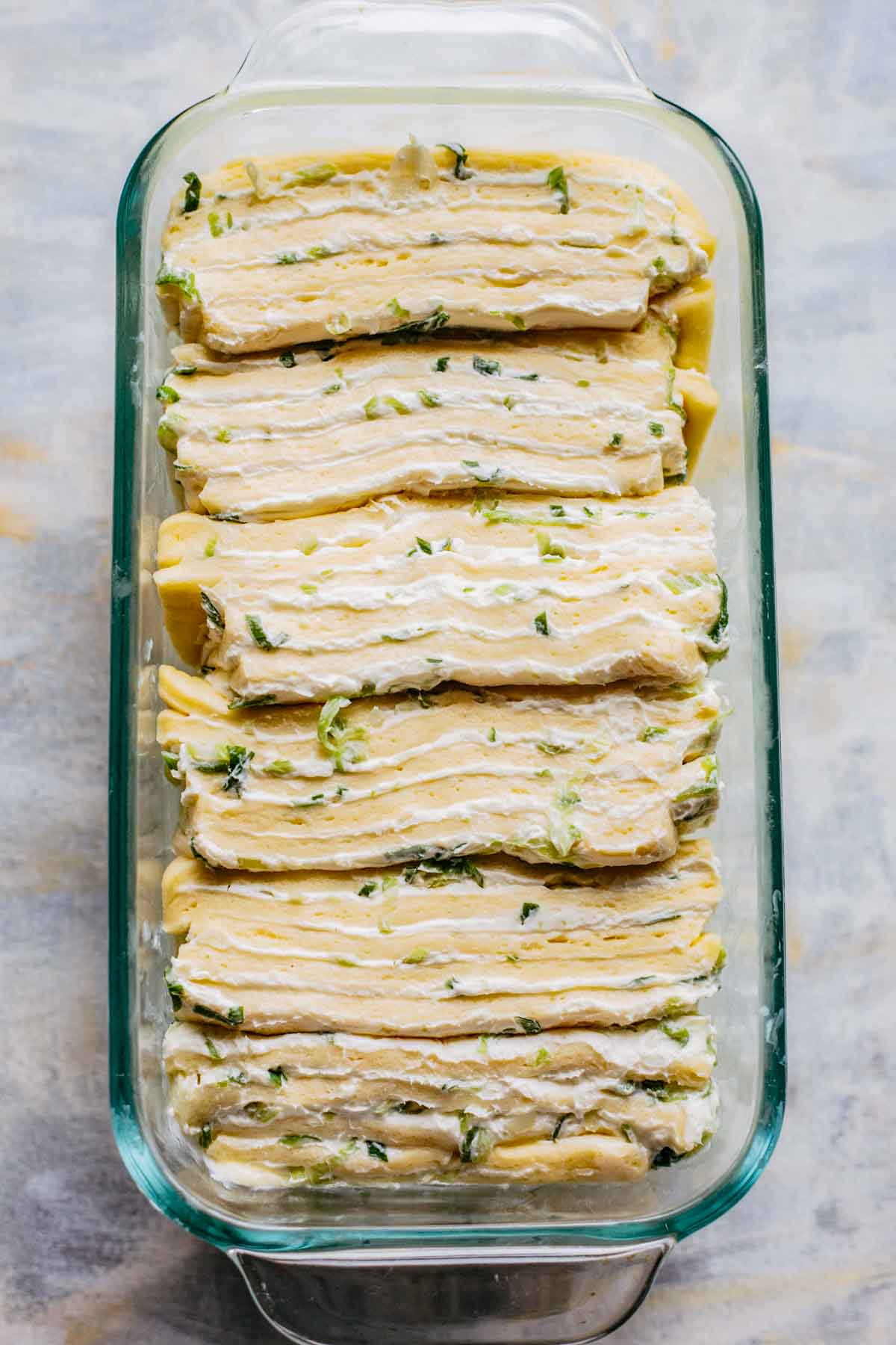 Overhead shot of stacked dough pieces with scallion cream cheese placed neatly in a glass loaf pan.