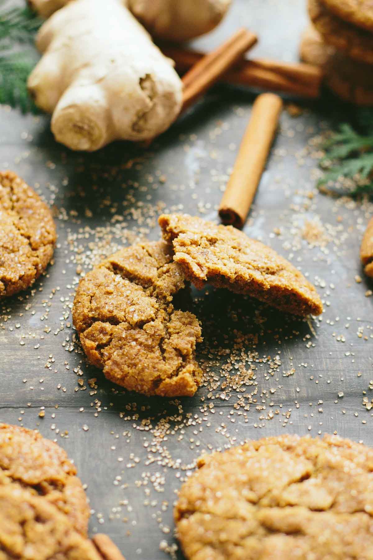 Close-up of a ginger molasses cookie broken in half showing its chewy interior, surrounded by ginger root and cinnamon sticks.