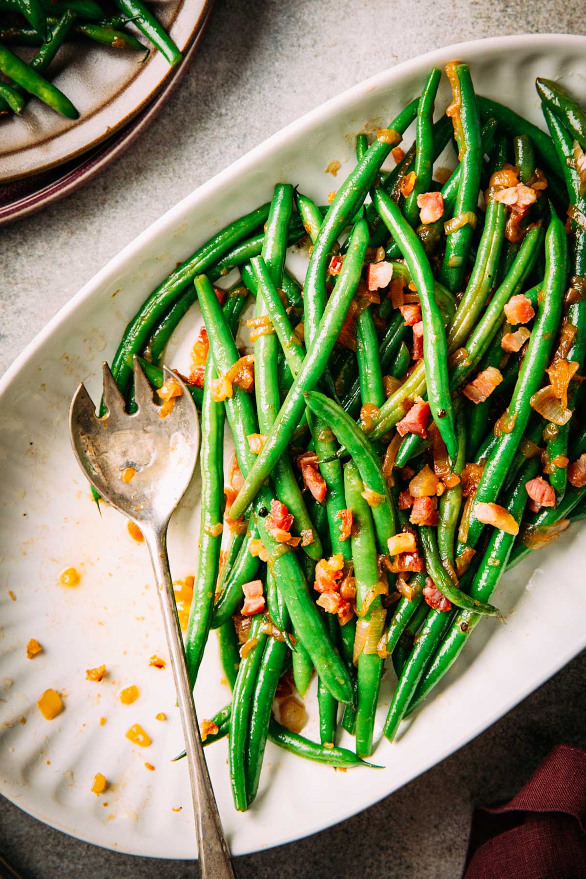 Overhead view of green beans with pancetta and shallots served on a white oval platter with a fork.