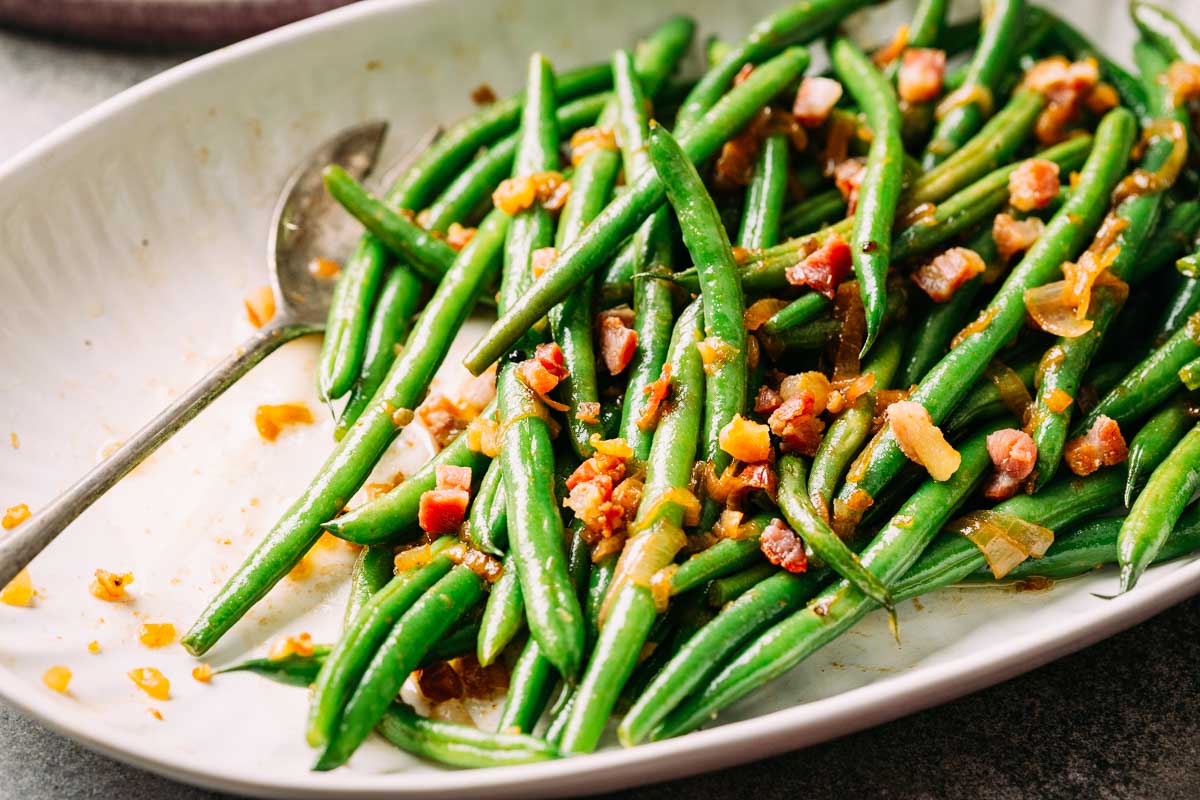 Angled view of green beans with pancetta on a white platter, showing their vibrant color and texture.