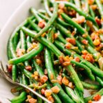 Close-up of glossy green beans topped with crispy pancetta pieces on a white platter.