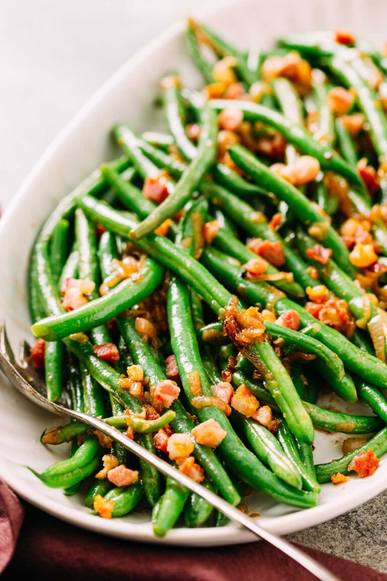 Close-up of glossy green beans topped with crispy pancetta pieces on a white platter.